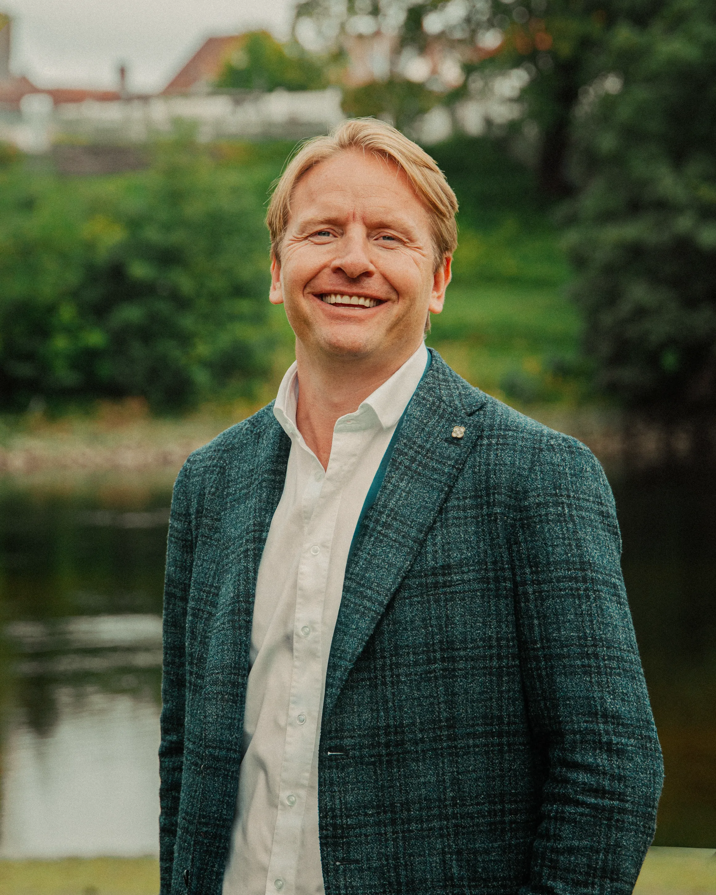 Smilende mann i rutet dressjakke fotografert utendørs ved vann